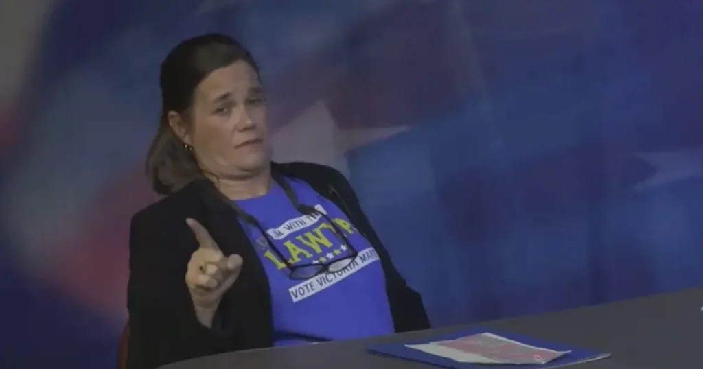 A woman wearing a blue shirt with campaign slogans is speaking at a podium.