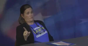 A woman wearing a blue shirt with campaign slogans is speaking at a podium.
