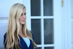 A woman with long blonde hair stands in front of a white door, smiling slightly.