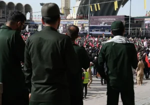 The photo depicts a large gathering of people, many wearing green uniforms, in what appears to be a public square or mosque, with flags and banners visible in the background.