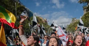 A vibrant crowd celebrates outdoors, waving flags and cheering under a clear blue sky.
