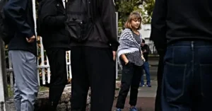A young girl in a striped shirt stands on a sidewalk, surrounded by adults, while another person checks their phone in the background.