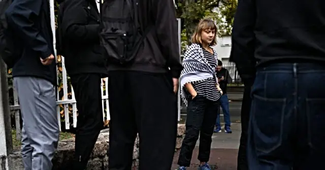 A young girl in a striped shirt stands on a sidewalk, surrounded by adults, while another person checks their phone in the background.