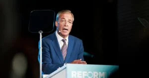 A man in a blue suit stands at a podium with the word "REFORM" visible, speaking at what appears to be a formal event.