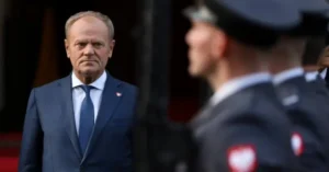 The image shows a man in a suit standing in front of a group of uniformed personnel, likely in a formal or official setting.