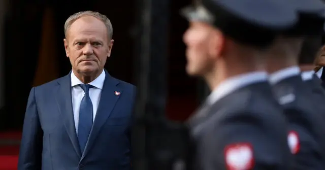 The image shows a man in a suit standing in front of a group of uniformed personnel, likely in a formal or official setting.