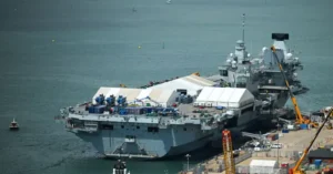 A large naval ship is docked at a port, with a white tent structure on its deck and construction equipment nearby.