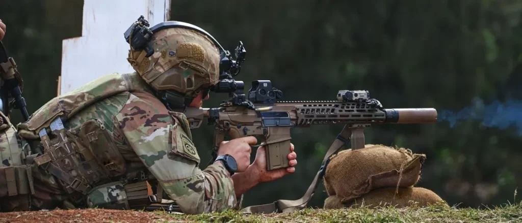 A soldier in camouflage gear is kneeling and aiming a rifle during a training exercise.