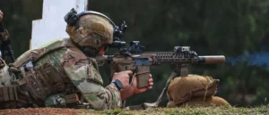 A soldier in camouflage gear is kneeling and aiming a rifle during a training exercise.