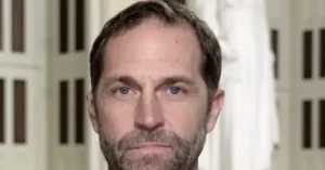 The image shows a close-up of a man with short brown hair and a beard, set against a background of vertical bars, possibly indicating a setting like a prison or detention facility.