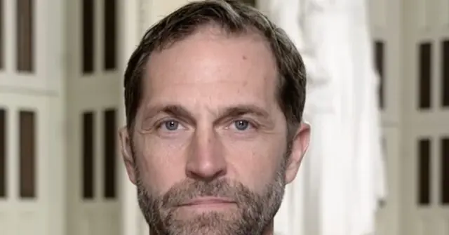 The image shows a close-up of a man with short brown hair and a beard, set against a background of vertical bars, possibly indicating a setting like a prison or detention facility.