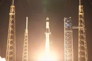 A rocket lifts off from a launchpad, surrounded by support structures, with bright flames and smoke visible beneath it.