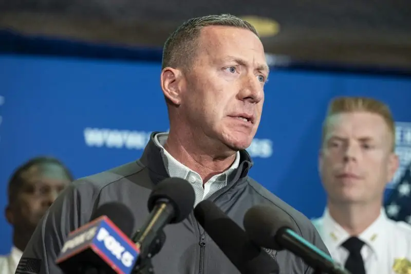 A man speaks at a press conference with microphones in front of him, while two other individuals stand in the background.