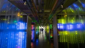 The image depicts a high-tech data center with rows of servers illuminated by colorful lights, and two individuals walking through the corridor.