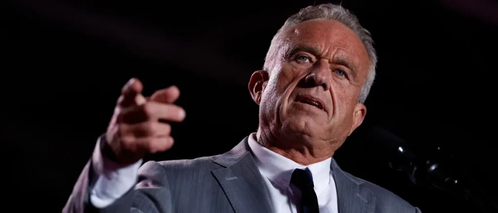 The image shows a man in a suit gesturing with his right hand while speaking, likely during a formal or political event.