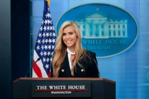 A woman stands at a podium labeled "The White House, Washington" with an American flag and the White House seal in the background.