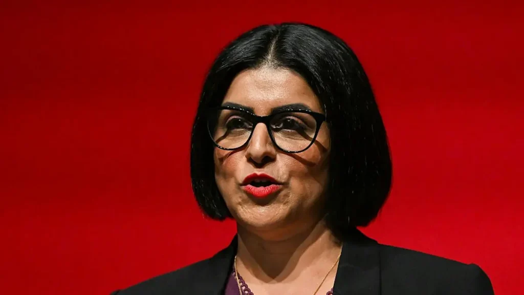 The image shows a person wearing glasses and a black blazer, standing against a red background.