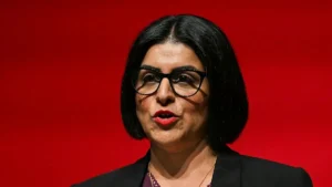 The image shows a person wearing glasses and a black blazer, standing against a red background.