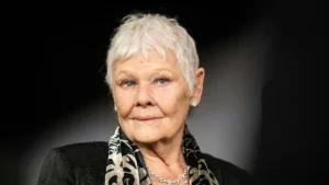 The image shows a person with short, white hair, wearing a black outfit with a patterned scarf and a necklace, set against a dark background.