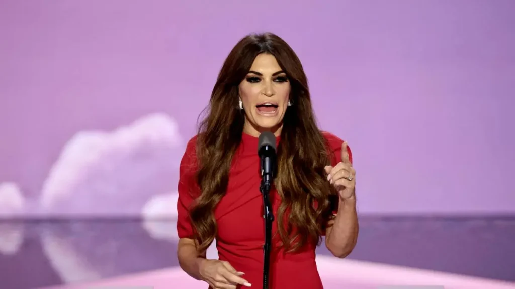 A woman in a red dress is giving a speech on stage, gesturing with her right hand.