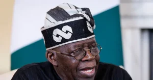 The image shows a person wearing a traditional headwrap with a distinctive pattern, speaking in what appears to be a formal or professional setting.