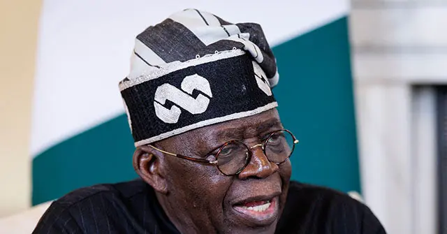 The image shows a person wearing a traditional headwrap with a distinctive pattern, speaking in what appears to be a formal or professional setting.