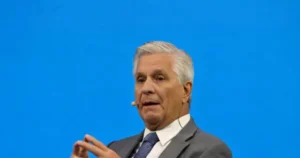 The image shows a man in a suit and tie, speaking with a microphone, against a blue background.