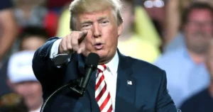 The image shows a person in a suit and tie speaking into a microphone, pointing towards the audience.