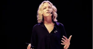 A woman is giving a presentation on stage, gesturing with her hands while holding a device in one hand.