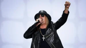 The image shows a performer on stage, wearing a black leather jacket, a cross necklace, and a black hat, holding a microphone and raising one fist in the air.