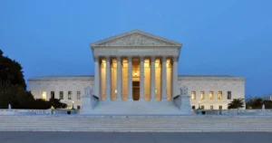The image shows the United States Supreme Court building illuminated at dusk, showcasing its iconic neoclassical architecture with columns and steps.