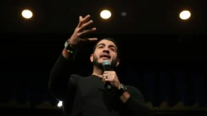 A man is speaking into a microphone while gesturing with his hand, likely during a public speaking event or performance.