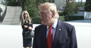 The image shows a man in a suit and red tie speaking outdoors, with a woman in a business suit standing behind him.