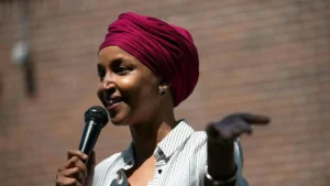A woman wearing a red turban speaks into a microphone while gesturing with her hand.