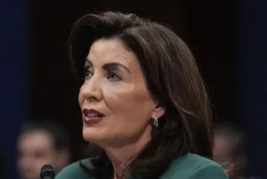 The image shows a woman speaking at a formal event, possibly a hearing or a press conference, as indicated by the microphone and the setting.
