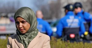 A woman wearing a hijab stands in the foreground while police officers on motorcycles are visible in the background.