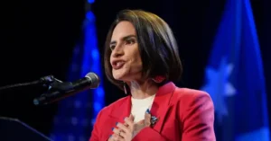 A woman in a red blazer is speaking at a podium with a microphone, with flags in the background.
