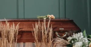 The image shows a wooden casket adorned with flowers and dried grasses, set against a dark green background.