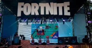 The image shows a stage setup for a Fortnite event, featuring a large screen displaying a map and various characters, with a "FAN FESTIVAL" banner and a stage setup for a presentation or performance.