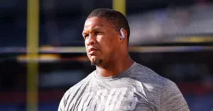 The image shows a person wearing a gray athletic shirt and earbuds, standing on a football field with a stadium in the background.