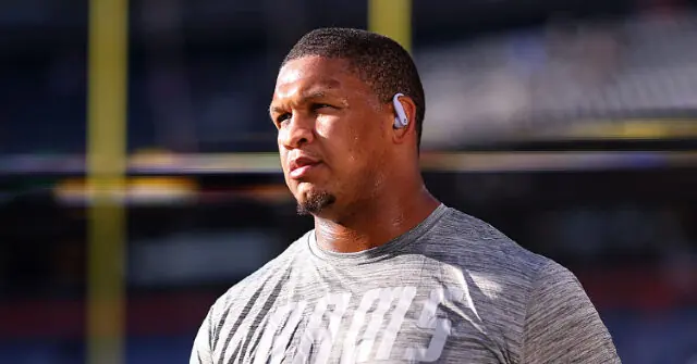 The image shows a person wearing a gray athletic shirt and earbuds, standing on a football field with a stadium in the background.