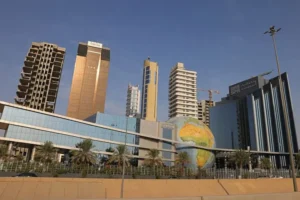 The image shows a modern urban landscape with tall buildings, a large globe sculpture, and palm trees, under a clear blue sky.