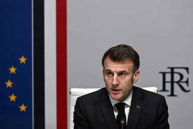 The image shows a man in a suit seated at a desk with a microphone, with the French flag and the European Union flag in the background.