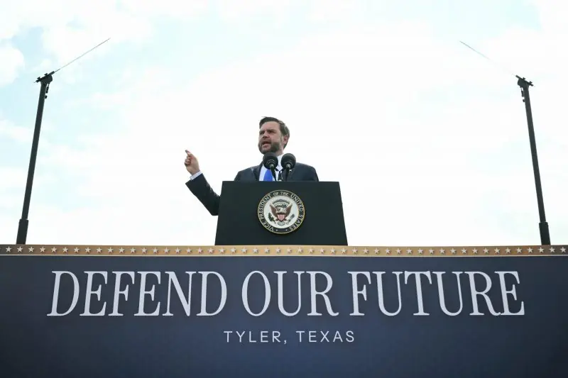 A man stands at a podium in Tyler, Texas, delivering a speech with the backdrop of wind turbines and a sign that reads "DEFEND OUR FUTURE."