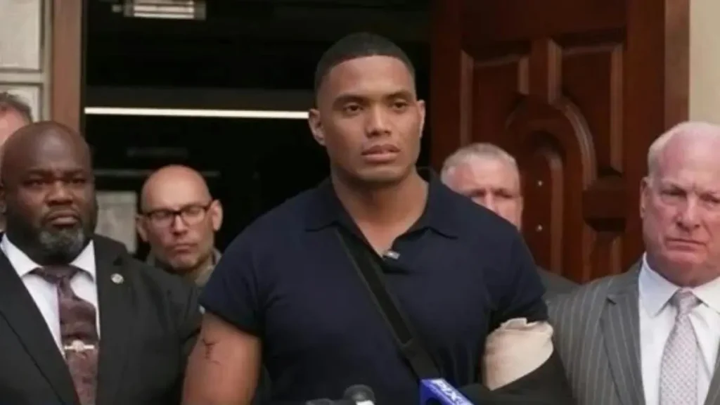 A man in a black shirt stands in front of a group of people, possibly during a press conference or public statement.