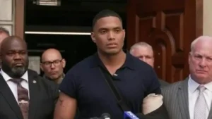 A man in a black shirt stands in front of a group of people, possibly during a press conference or public statement.