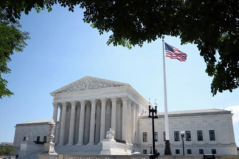 The image shows the United States Supreme Court building with a flag flying at half-staff, framed by trees and a clear blue sky.