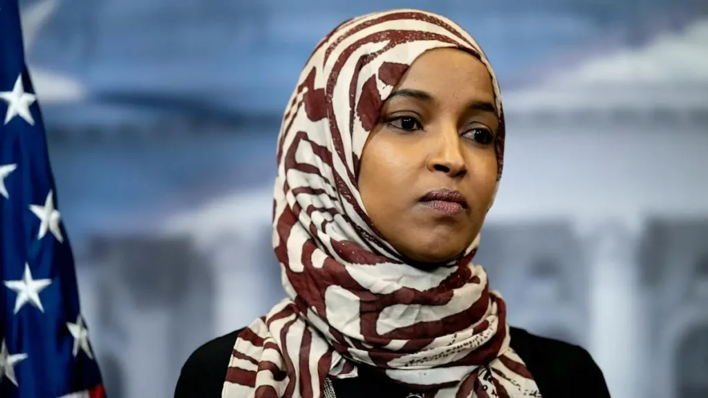 The image shows a person wearing a patterned hijab, standing in front of an American flag, suggesting a formal or political setting.