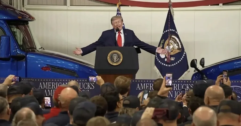 The image shows a political figure speaking at a podium with an American flag and presidential seal in the background, addressing a crowd of people.