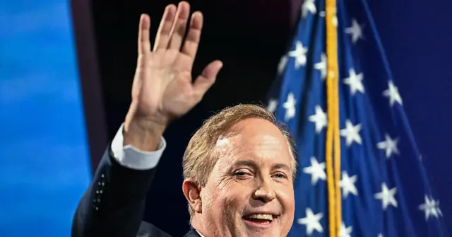 A man in a suit waves to the audience while standing in front of an American flag.